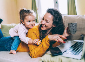 A white mother and daughter play on the couch while mom works on a laptop